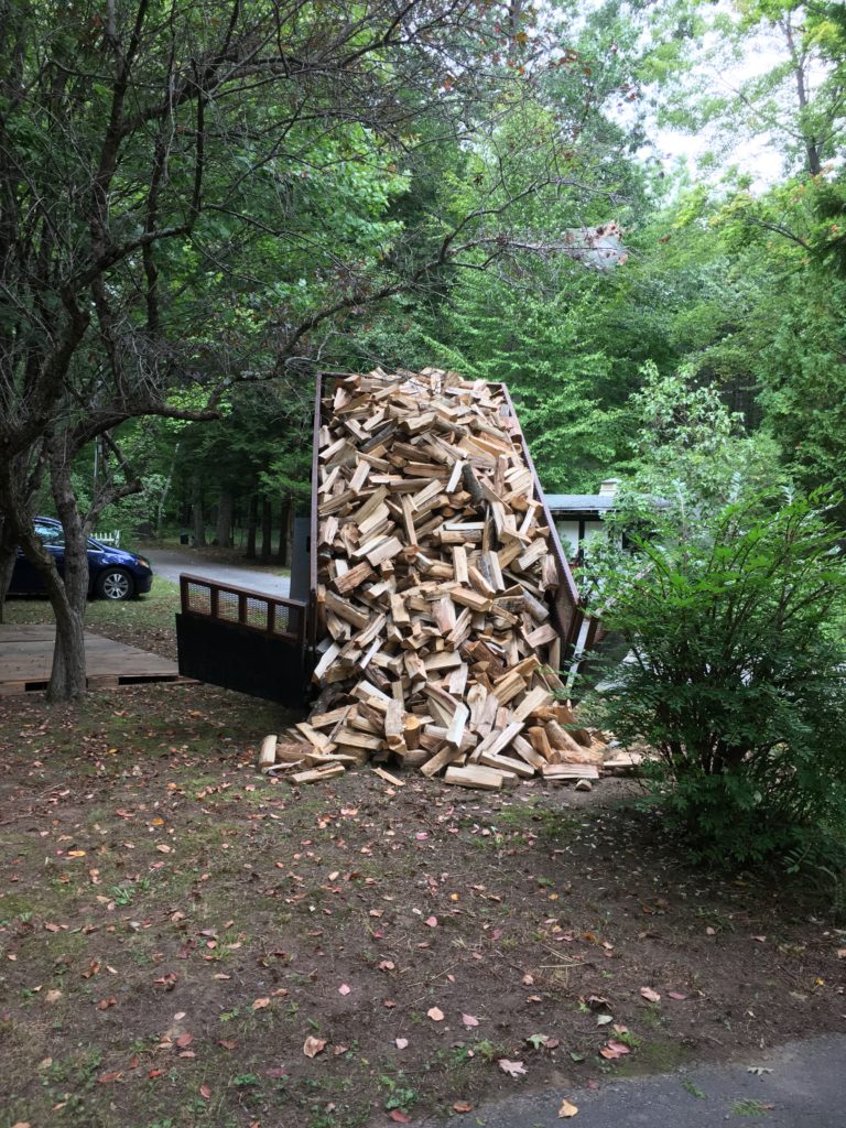 Two cords of firewood being dumped out of a dump trailer behind a pickup truck into the yard next to where the wood is to be stacked.