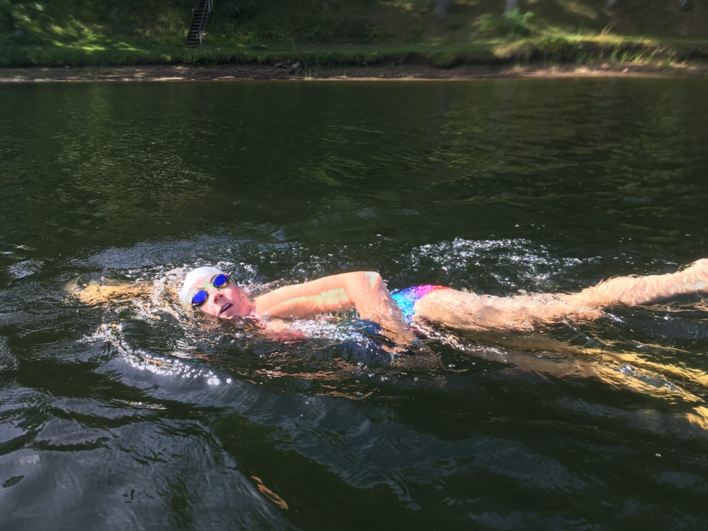 Kelsey swimming in the Hudson River with a swim cap and goggles with the shoreline in the background.