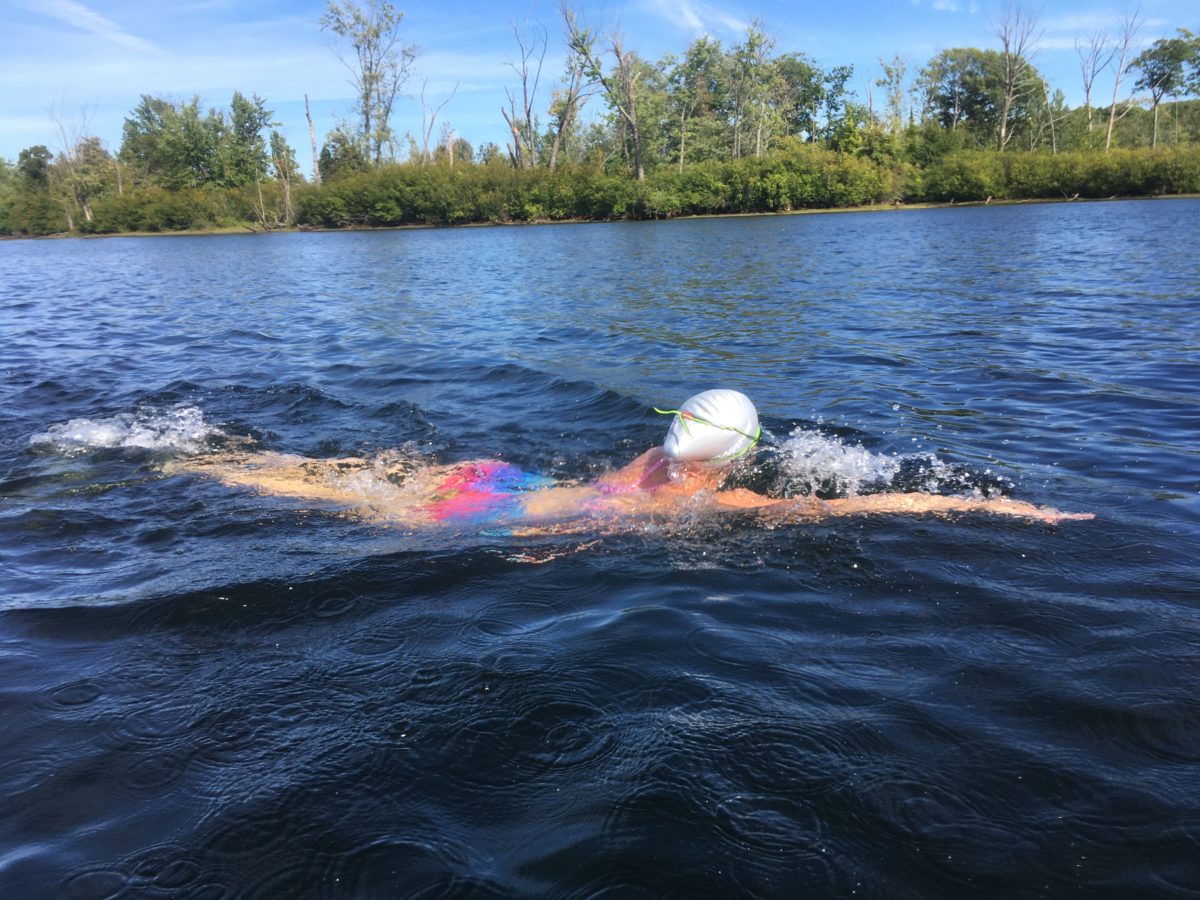 Kelsey in a swim cap and goggles swimming in the Hudson River with a tree-covered island in the background.