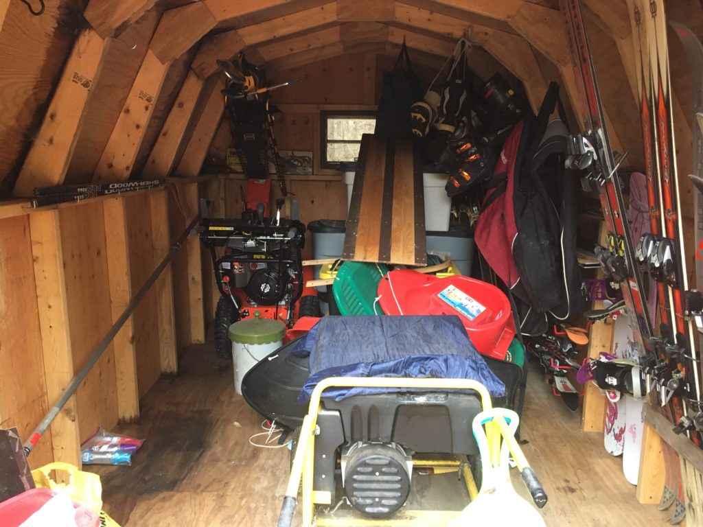 A photograph of the inside of our storage shed containing our winter gear, including skis, snowboards, the snowblower, a generator, sleds, and several tubs.