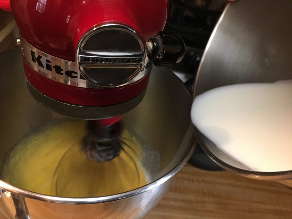 A photo of hot milk being slowly aded to the egg/milk mixture in the Kitchen Aid mixer while the whisk attachment is running.