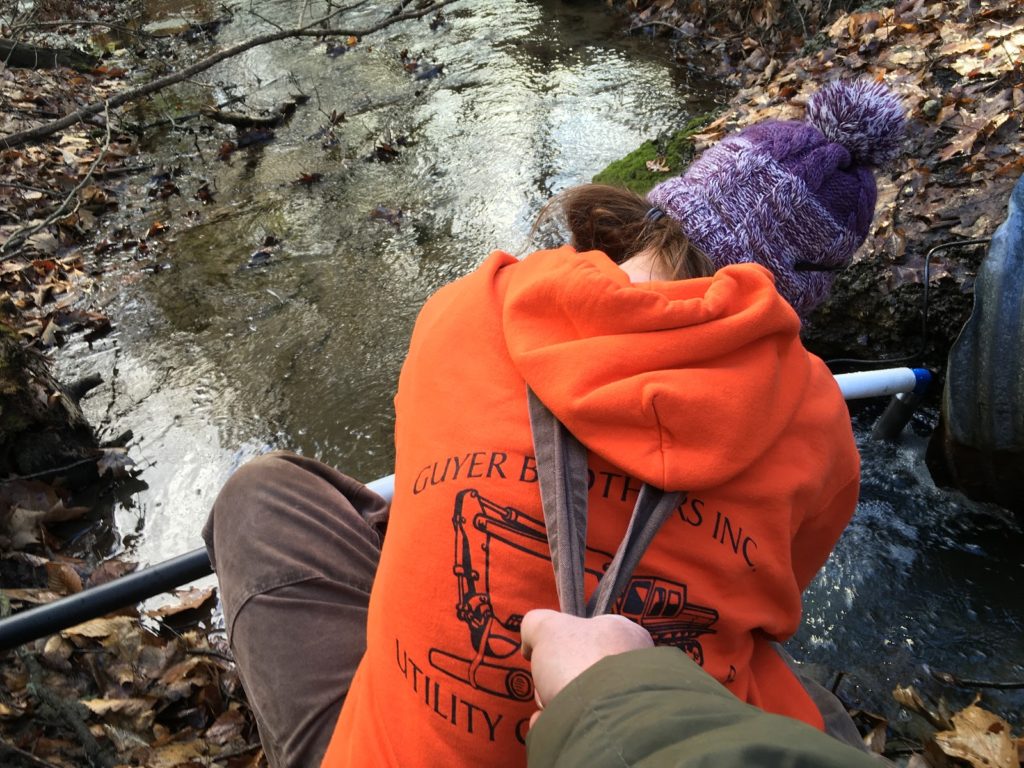 A photo of Kevin holding Kelsey above the stream by her overall straps while she installs the sump pump.