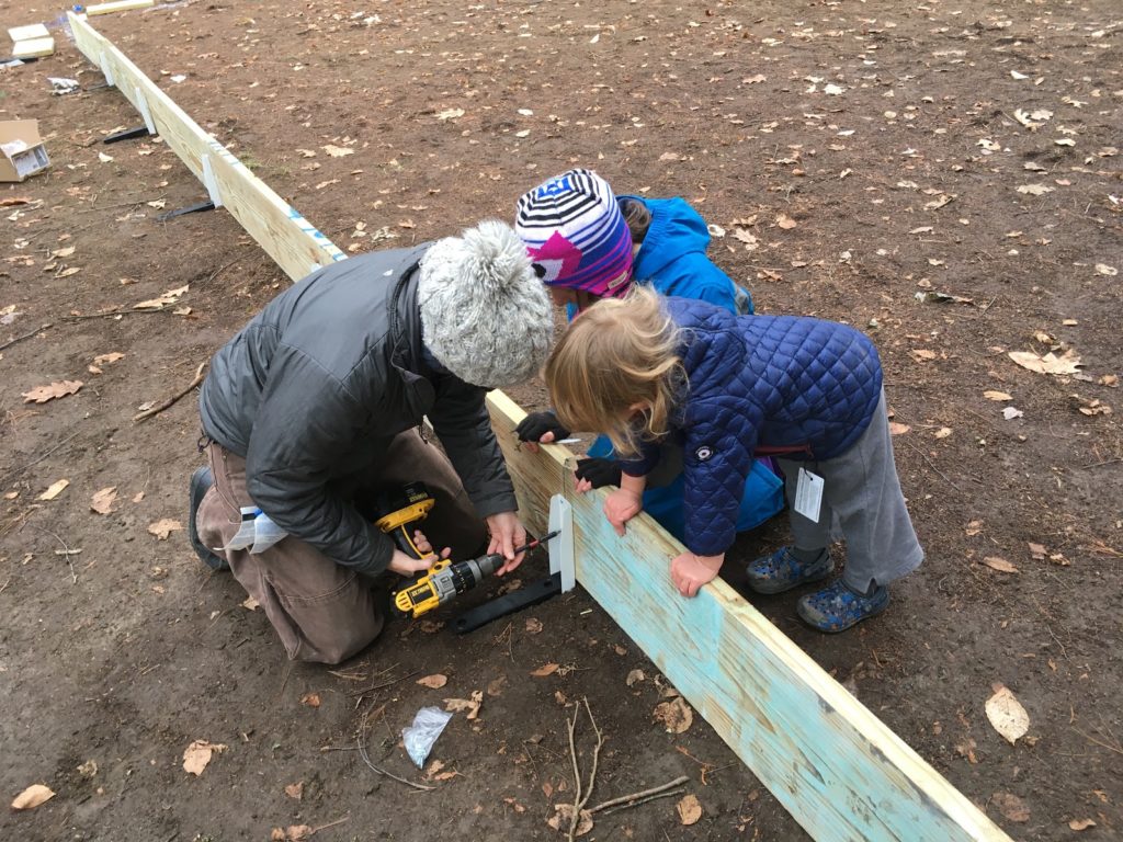 A photo of Kelsey screwing in a support stake to the rink wall while Rayleigh and Grayson help support the boards.