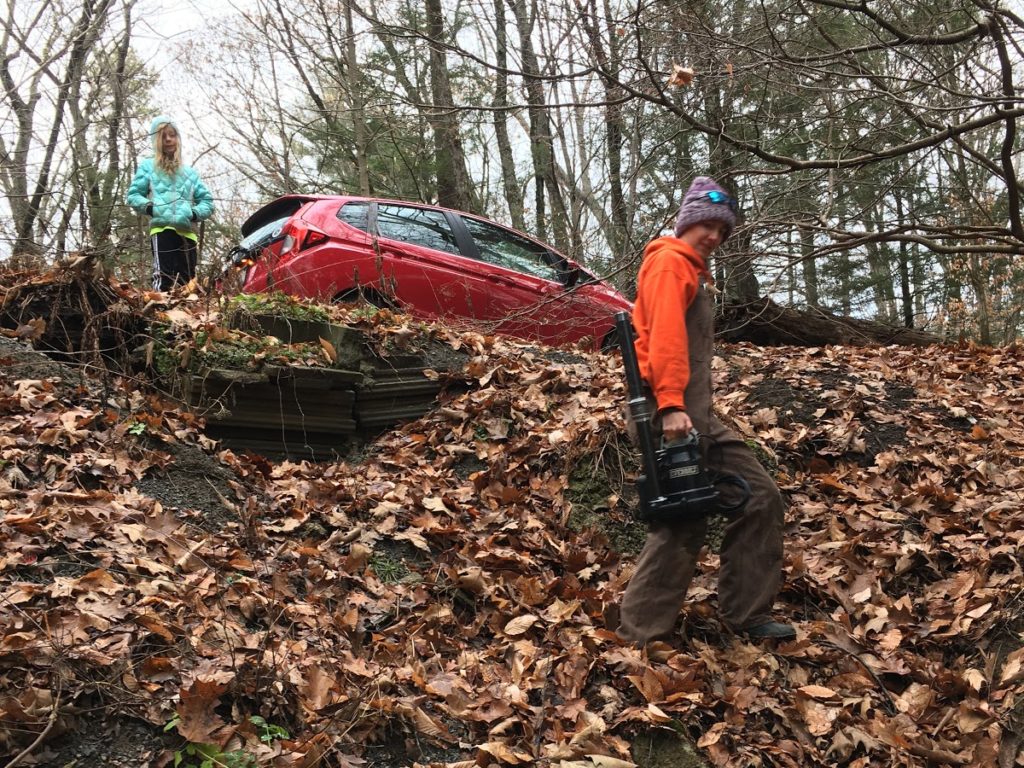 A photo of Kelsey in overalls carrying a sump pump down the bank to the stream while Dillon looks on.