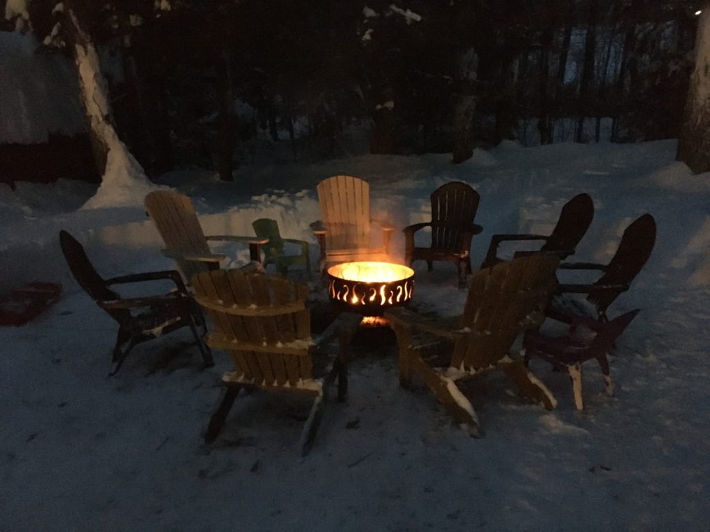 A photo of a fire pit in a snowy yard surrounded by Adirondack chairs.