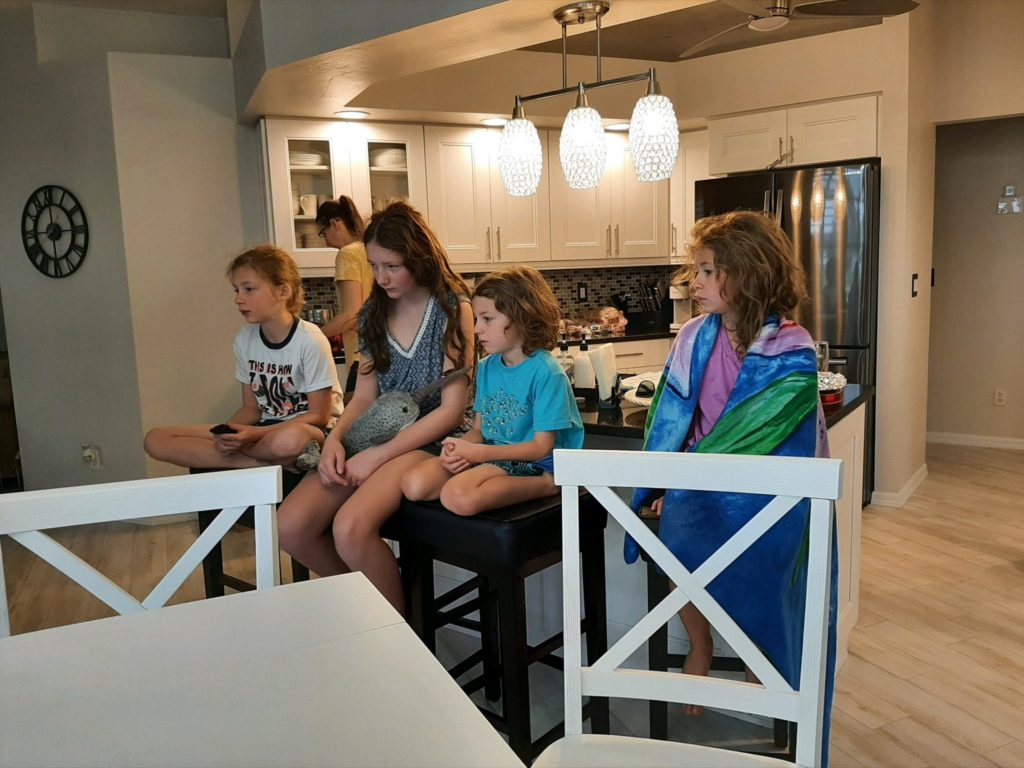 A photo of Dillon, Rayleigh, Grayson, and Ainsley watching TV in their pajamas while Kelsey works in the kitchen