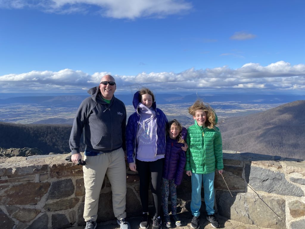 A photo of Kevin, Rayleigh, Ainsley, and Dillon being buffeted by the string winds at the top of the highest peak in Shenandoah National Park