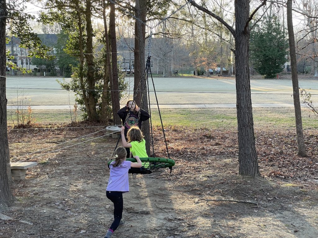 A photo of Rayleigh pushing Grayson on a monkey swing while Evie looks on