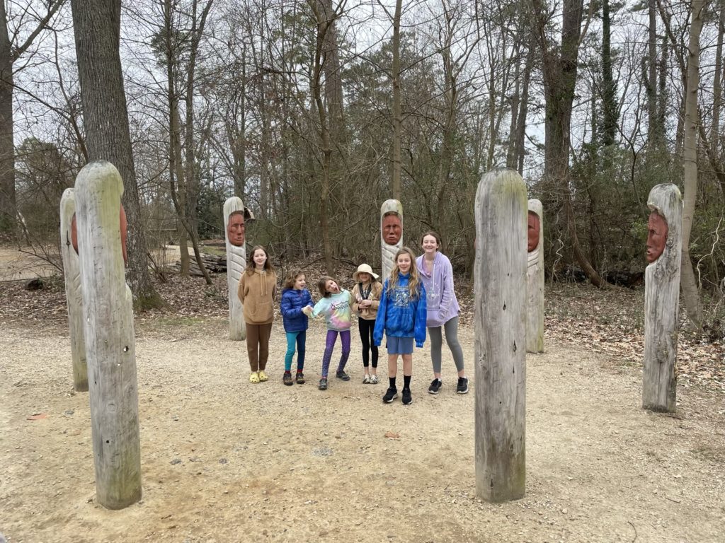 A photo of Evie, Grayson, Ainsley, Olivia, Dillon, and Rayleigh standing in the middle of a circle of seven posts with faces pointed inward