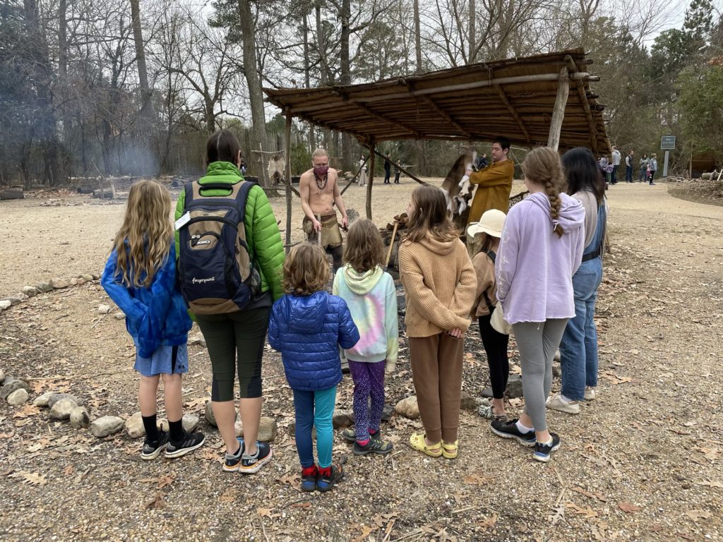 A photo of Dillon, Kelsey, Grayson, Ainsley, Evie, Olivia, Rayleigh, and Emily watching a Powhatan cooking demonstration in an open air kitchen