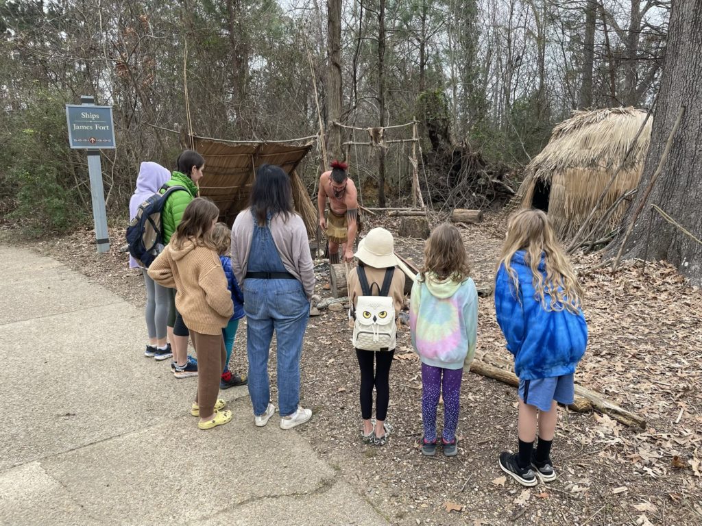 A photo of Rayleigh, Kelsey, Grayson, Evie, Emily, Olivia, Ainsley, and Dillon listening to a man dressed in 17th century Powhatan clothing describe the process of making bows