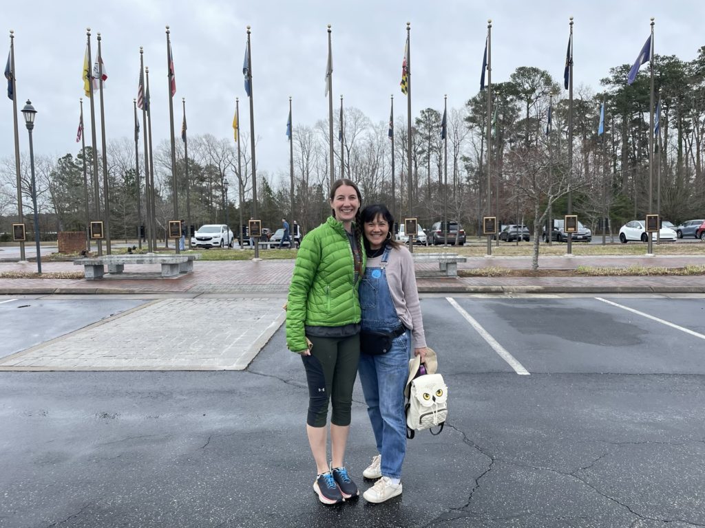 A photo of Kelsey and Emily in the parking lot at Jamestown with a series of flags in the background