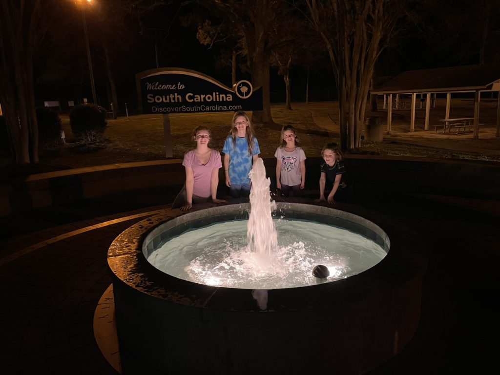 A photo of Rayleigh, Dillon, Ainsley, and Grayson standing behind a fountain with a Welcome to South Carolina sign in the background at night