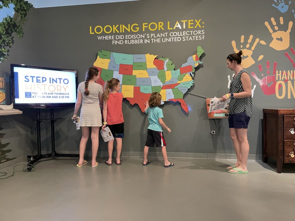 A photo of Rayleigh, Dillon, Grayson, and Kelsey looking at a display labeled "Looking for Latex: Where did Edison's plant collectors find rubber in the United States?"