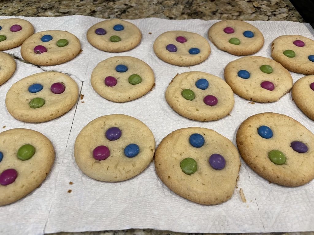 A photo of sugar cookies with colorful candies on them cooling on a paper towel