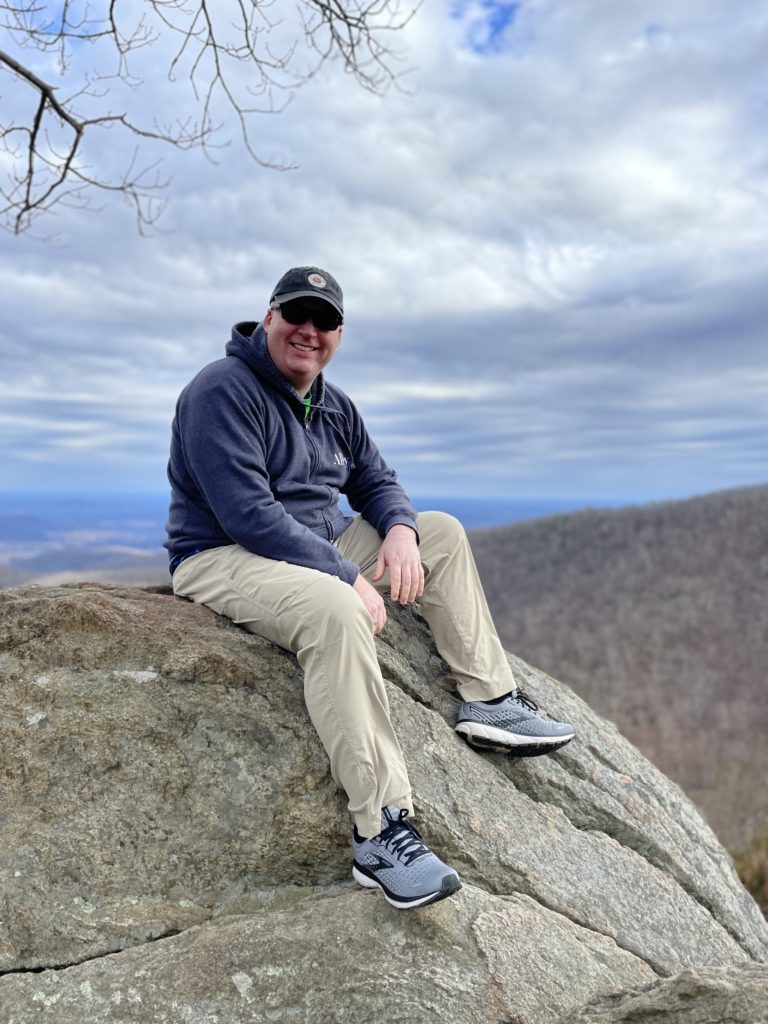 A photo of Kevin in a hat and sunglasses sitting on a large rock in Shenandoah National Park with the Appalachian mountains in the background