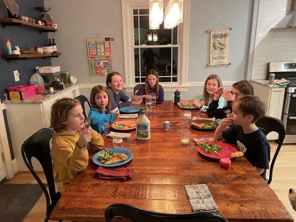 A photo of Grayson, Ainsley, Rayleigh, Mia, Dillon, Maisie, and Milo eating pizza and salad at a large wooden table