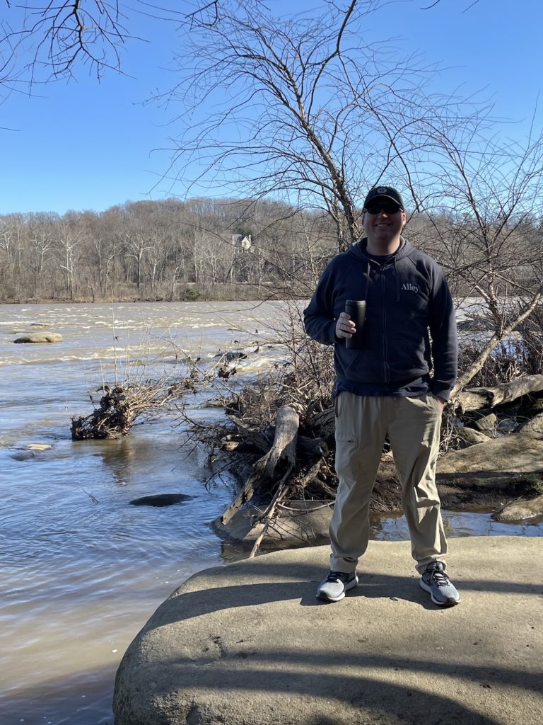 A photo of Kevin in hat and sunglasses holding a coffee standing on a rock in front of the James River
