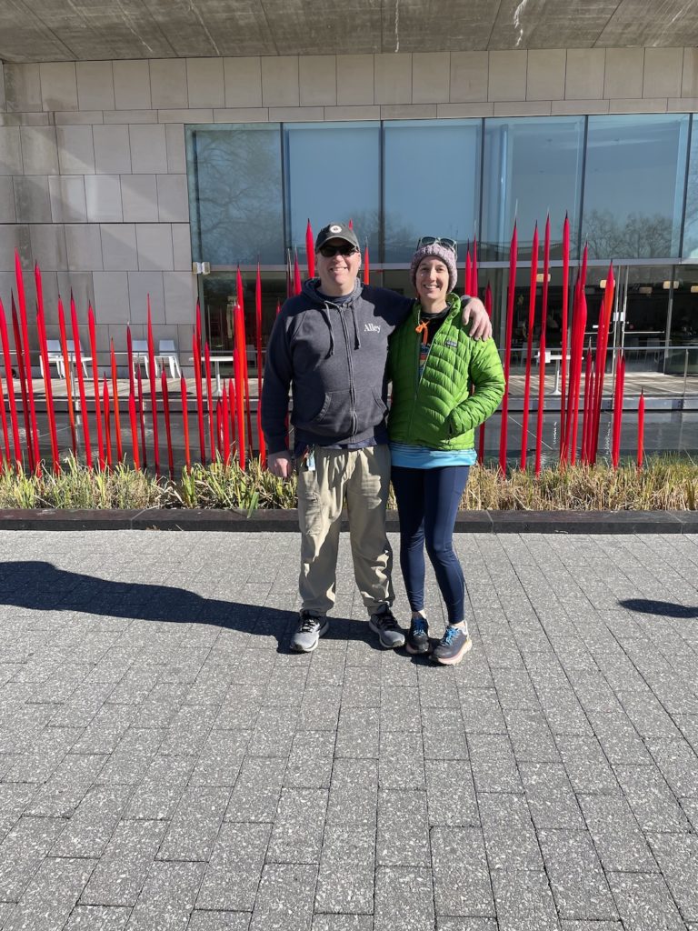 A photo of Kevin and Kelsey in front of an art installation of blown glass red reeds in water outside of the Virginia Museum of Fine Arts