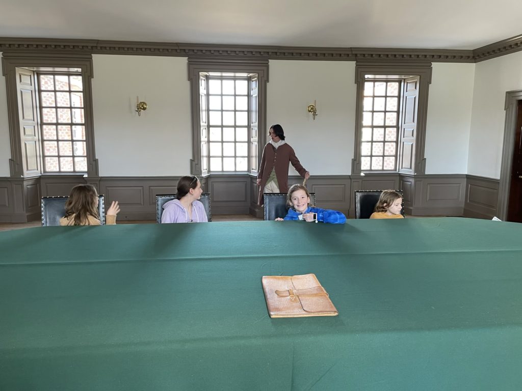 A photo of Evie, Rayleigh, Dillon, and Grayson listening to a speaker in the capitol at Colonial Williamsburg