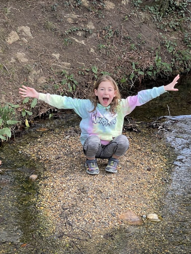 A photo of Ainsley doing a silly pose on a small island in the middle of a stream running through Colonial Williamsburg
