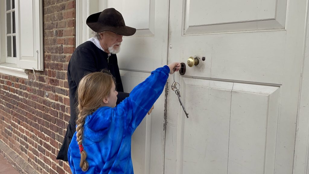 A photo of Dillon locking the courthouse using a giant key while a tour guide in period costume looks on in Colonial Williamsburg
