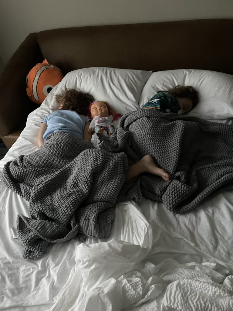 A photo of Ainsley and Grayson asleep in a hotel bed with a baby doll and an orange plush fish in bed with them