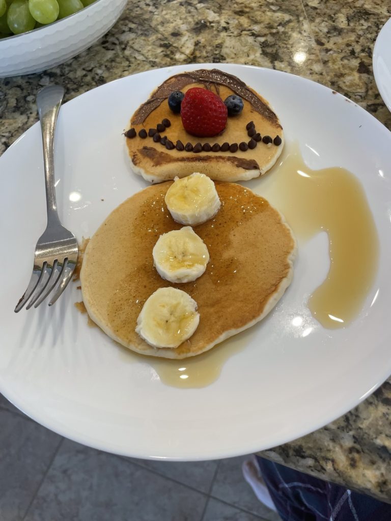 A photo of two pancakes decorated to look like a clown. The bottom pancake is the body and has three banana slices for buttons. The top pancake is the head and has Nutella hair, blueberry eyes, a strawberry nose, and a mouth made of chocolate chips.