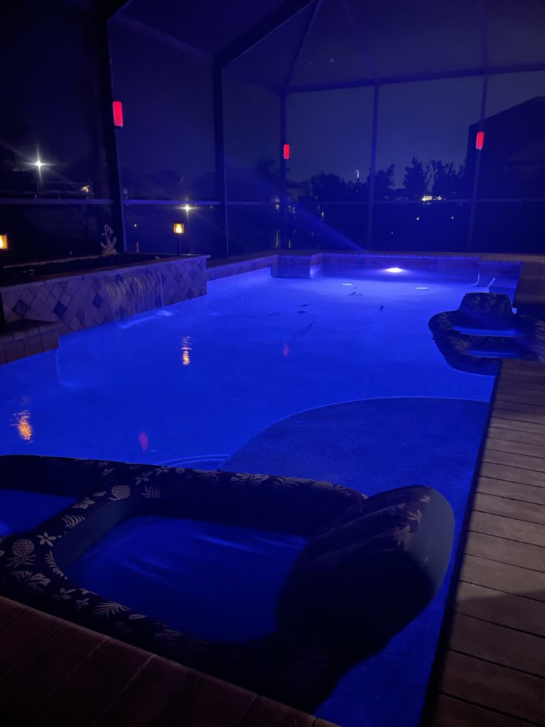 A photo of the pool at night, lit up by red lights on the posts of the screens and a violet light in the pool itself