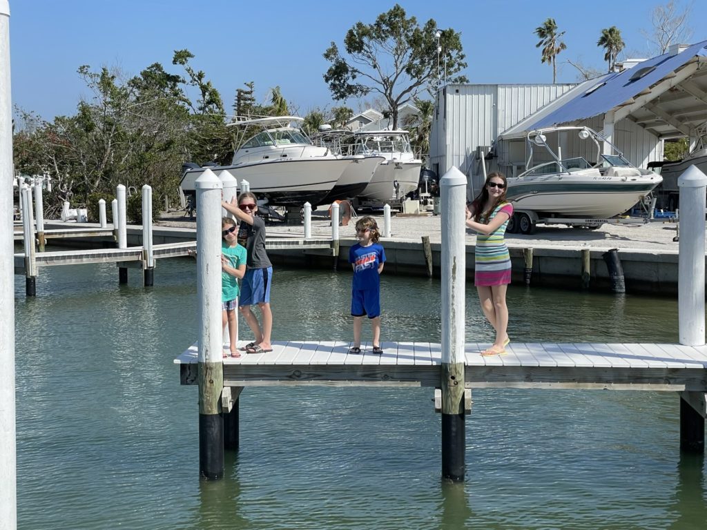 A photo of Ainsley, Dillon, Grayson, and Rayleigh in sunglasses standing on a pier with boats in the background on trailers.