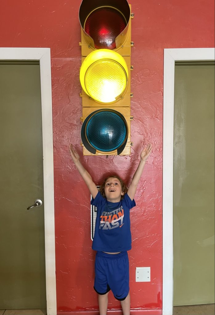A photo of Grayson standing under a four foot tall traffic light with the yellow light lit up and his hands in the air
