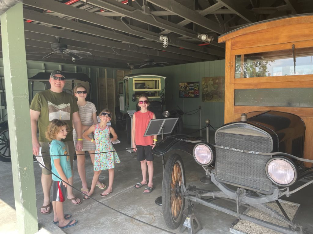 A photo of Kevin, Grayson, Rayleigh, Ainsley, and Dillon standing next to several early Ford automobiles