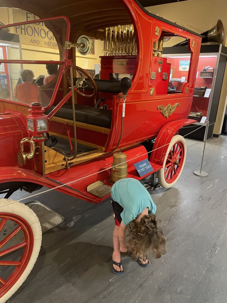 A photo of Grayson being shy and hiding from the camera in front of an early Ford automobile painted red with white tires with a calliope in the back, which is a sort of steam-powered organ with several dozen shiny pipes sticking out on top