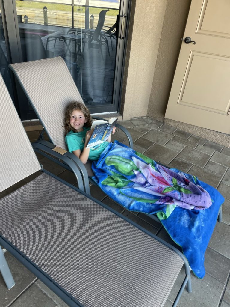 A photo of Ainsley reading a thick book on a reclining chair under a towel on the pool deck