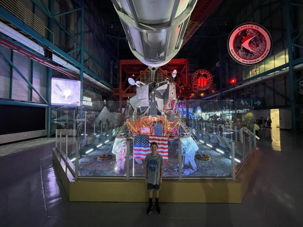 A photo of Dillon standing in front of a replica of the lunar lander with astronauts on a spacewalk planting the American flag