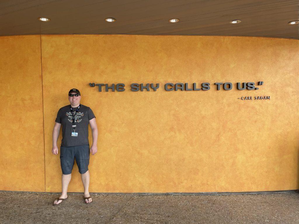A photo of Kevin standing in front of a wall with the text "The sky calls to us. - Carl Sagan" written on it
