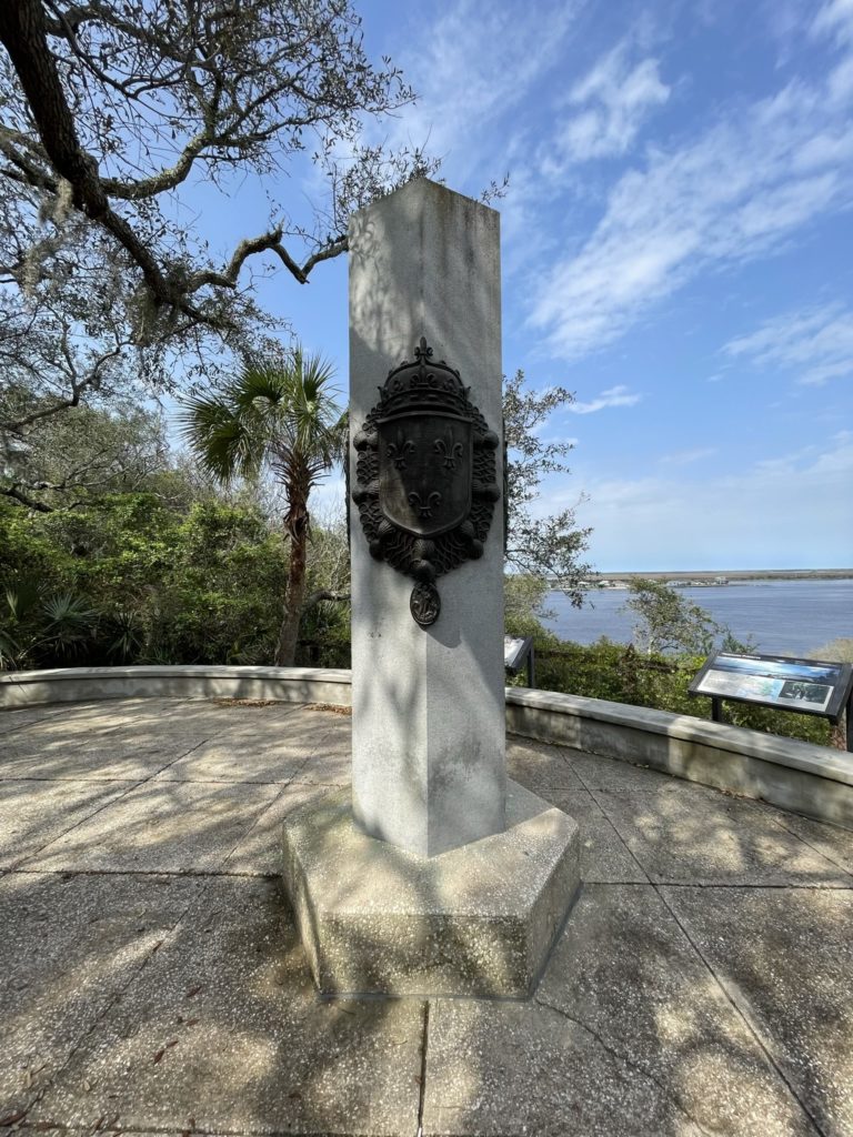 A photo of a stone pillar with a fleur de lis emblem on a metal plaque affixed to its front with a river in the background