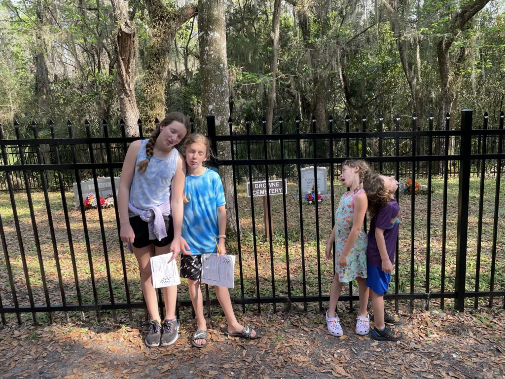 A photo of Rayleigh, Dillon, Ainsley, and Grayson standing in front of the fence around the Browne Cemetary and playing dead