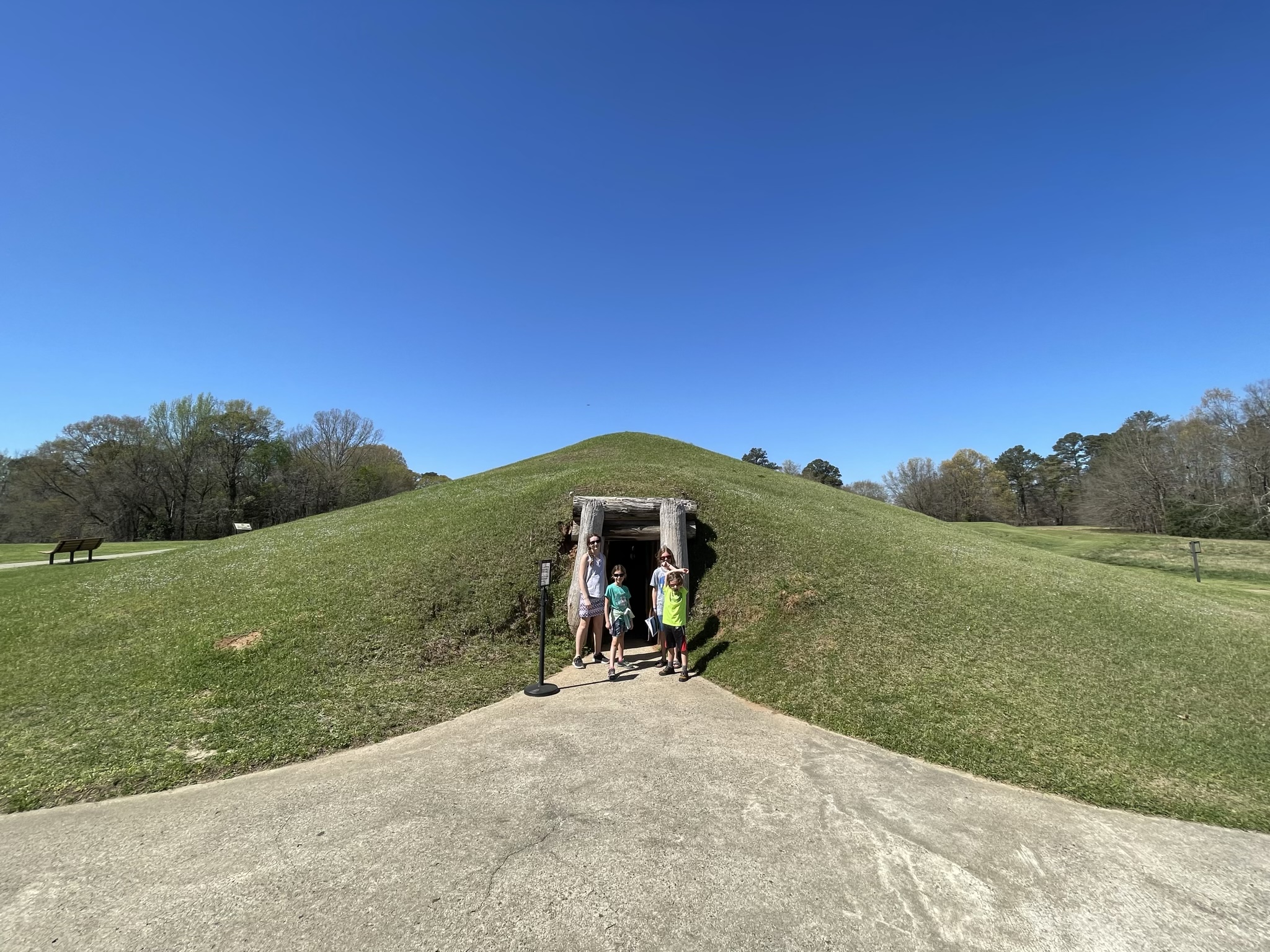 A photo of Rayleigh, Ainsley, Dillon, and Grayson standing in front of the wood framed entrance to an underground structure with a grassy mound forming its roof
