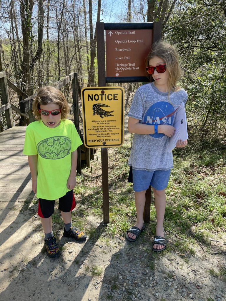 A photo of Grayson and Dillon making scared faces in reaction to a sign that says "Notice: alligators are common in this area. They can be dangerous and should not be approached, frightened or fed. Please give them the respect they deserve. Keep your distance."