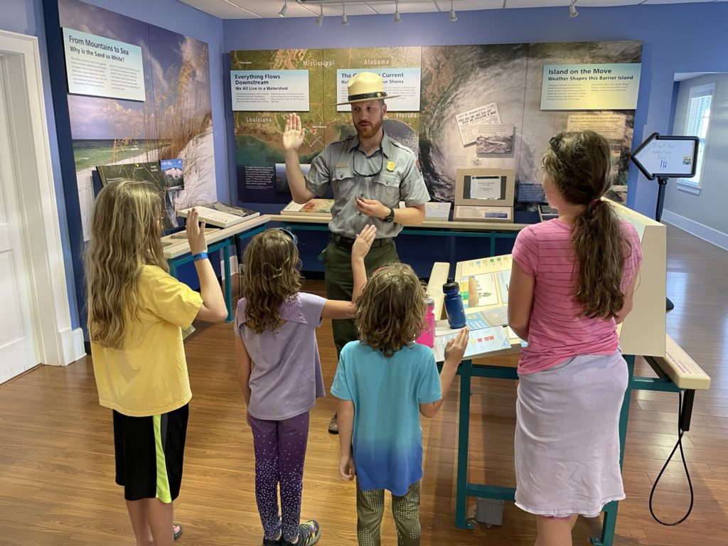 A photo of Dillon, Ainsley, Grayson, and Rayleigh taking the Junior Ranger pledge from a national park ranger inside of the ranger station