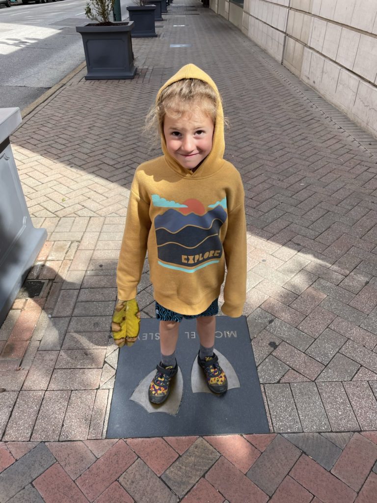 A photo of Grayson in a hoodie standing on a plaque in the sidewalk with duck feet on it