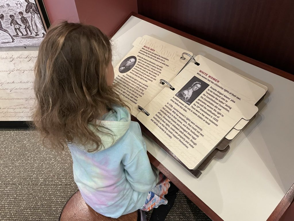 A photo of Ainsley reading a display about the rights different people would have at the nation's founding. The page she is on lists the rights for white men and white women.