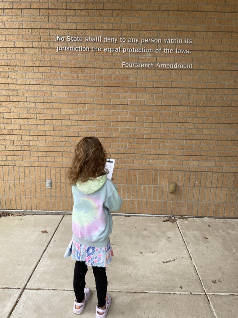 A photo of Ainsley standing in front of a quote on the wall of a brick building that says "[No State shall] deny to any person within its jurisdiction the equal protection of the laws. - Fourteenth Amendment"