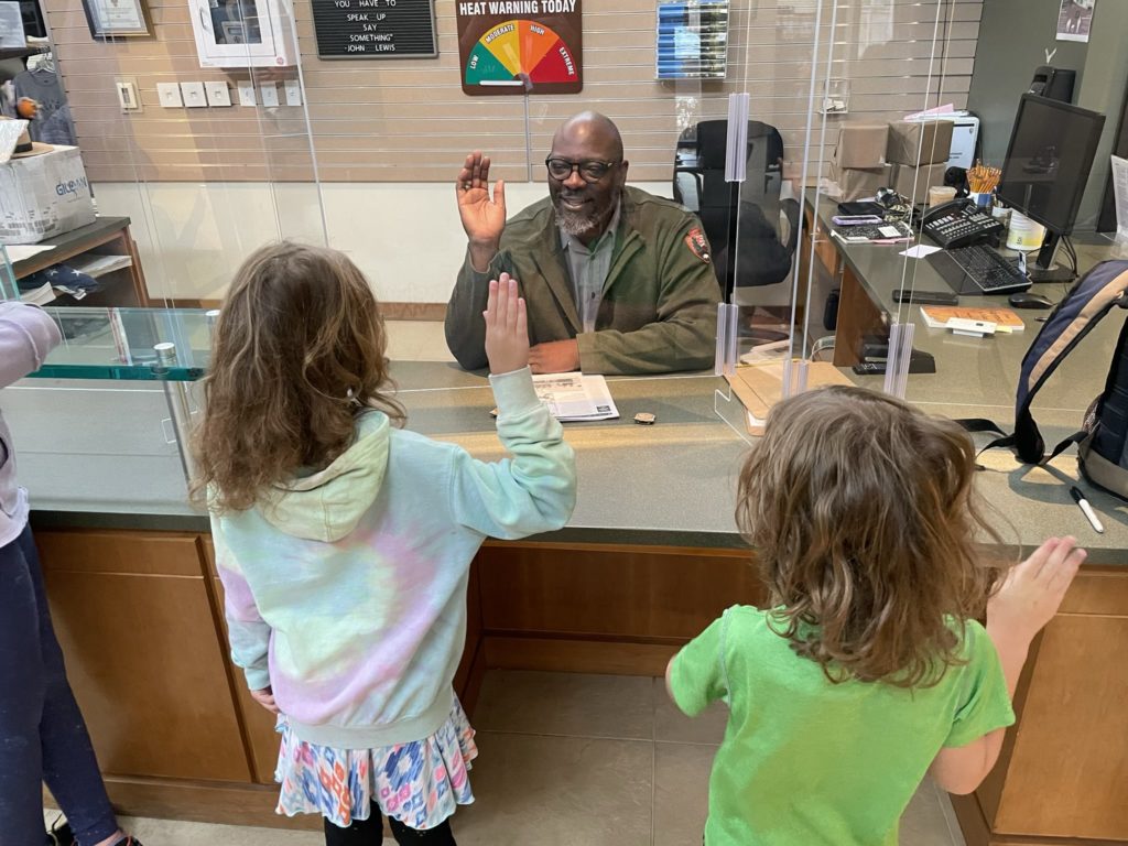 A photo of a Black national park ranger leading Ainsley and Grayson through the Junior Ranger pledge. All three have their right hands raised.