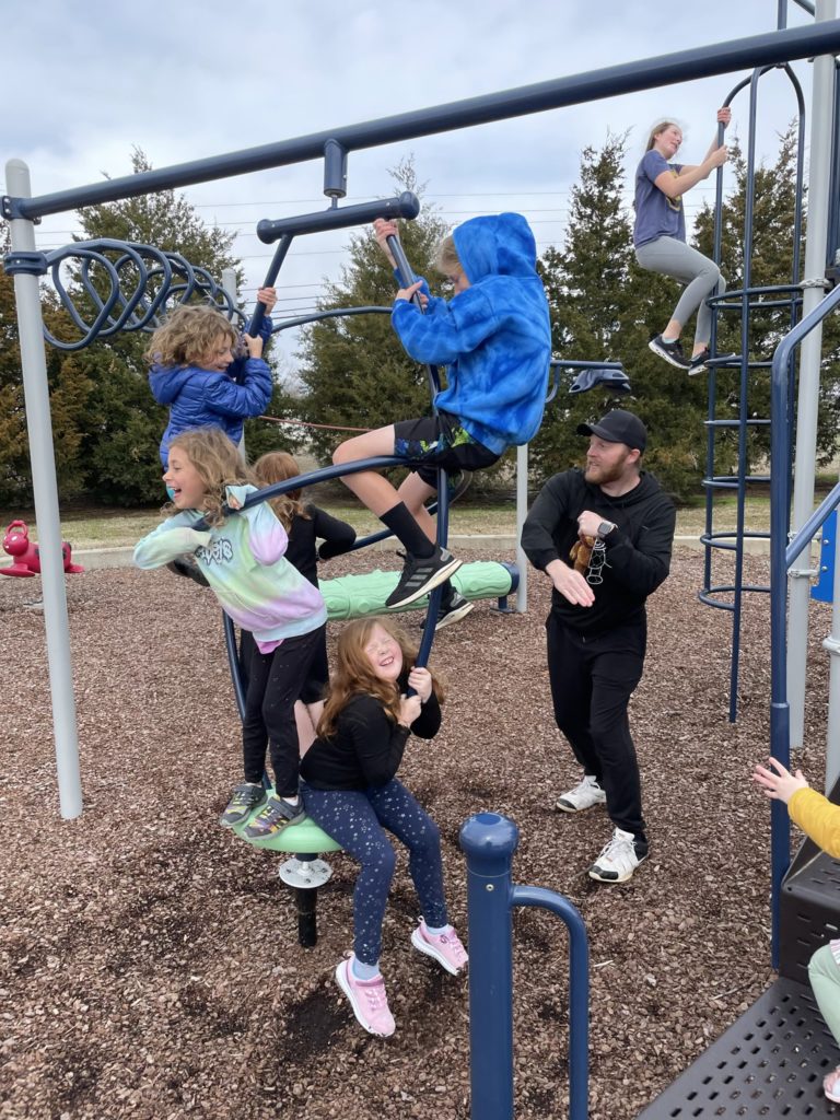 A photo of Kevin's cousin Tim spinning a piece of playground equipment with his daughters Hailey and Harper as well as Grayson, Ainsley, and Dillon on it, while Rayleigh climbs another playground structure in the background