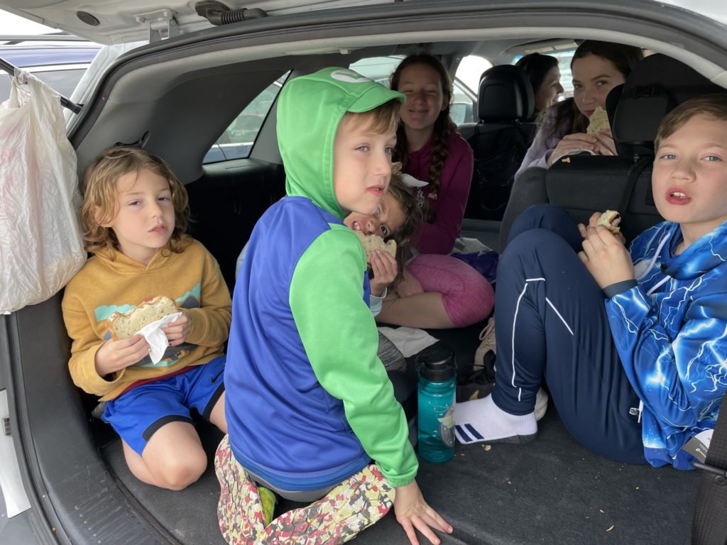 A photo of Grayson, Peter, Ainsley, Rayleigh, Myna, and Milo eating lunch in the back of Ashley's SUV