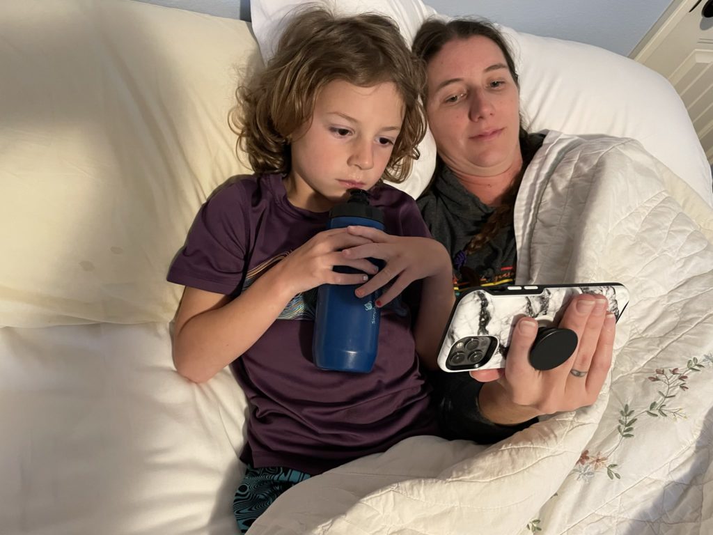 A photo of Grayson and Kelsey laying in bed watching a video on Kelsey's phone. Grayson is holding a water bottle near his mouth.