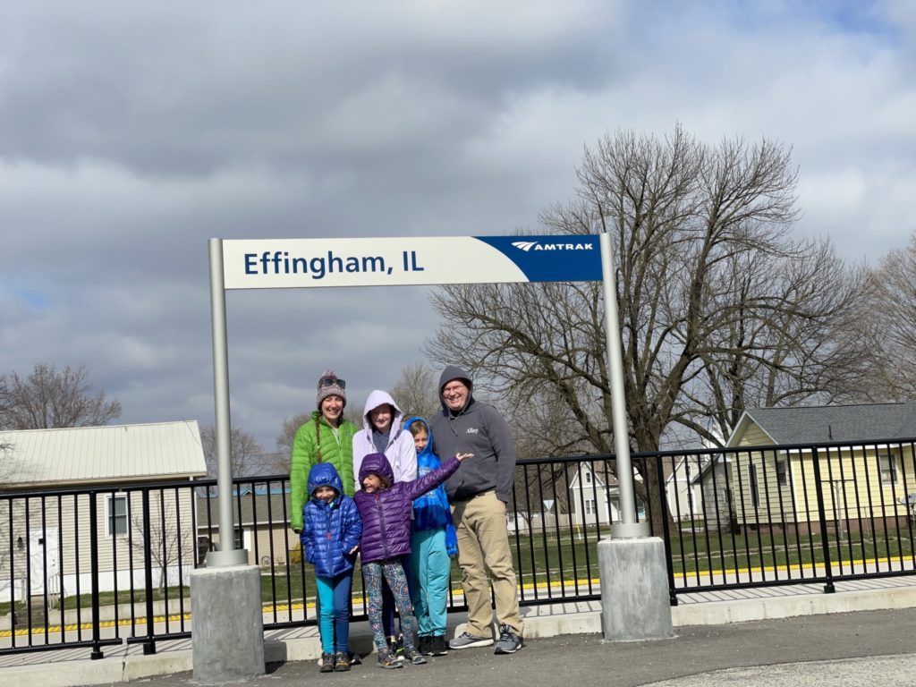 A photo of Grayson, Kelsey, Ainsley, Rayleigh, Dillon, and Kevin posing under a sign at an Amtrak station that says "Effingham, IL"