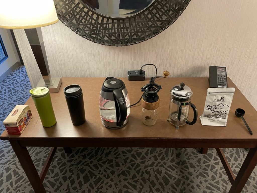 A photo of a box of tea, two travel mugs, an electric kettle, a hand grinder for coffee, a French press, an bag of coffee beans, and a scoop on a table in a hotel room